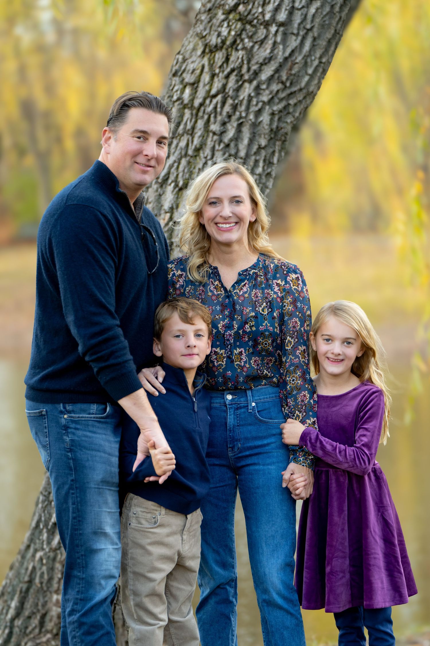 A family of four poses together for a portrait against a tree with autumn foliage in the background.