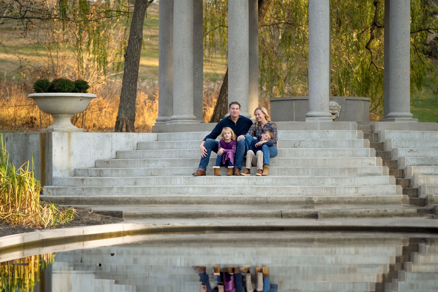 People sitting on stone steps near water with columns in a park during golden hour create a mirror reflection.