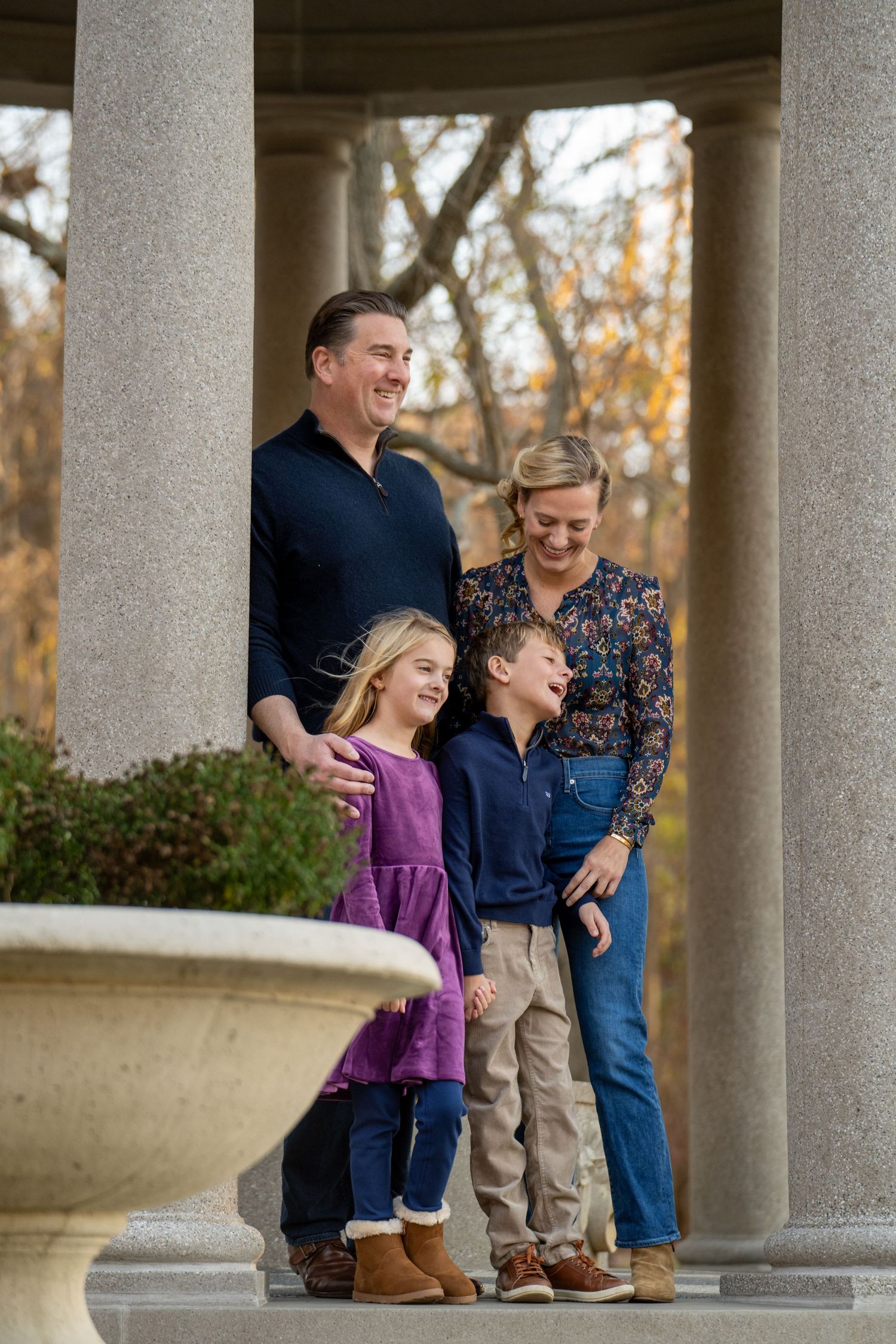 A family stands together in casual fall outfits beneath white stone pillars in an outdoor setting at sunset.
