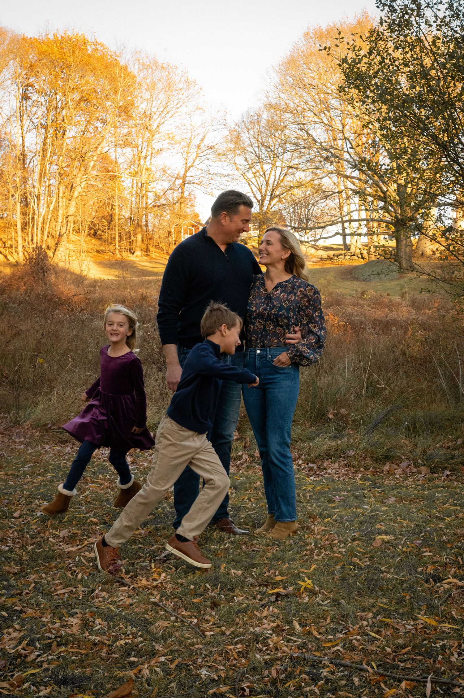 A family enjoys a playful moment together outdoors on an autumn afternoon with golden trees in the background.