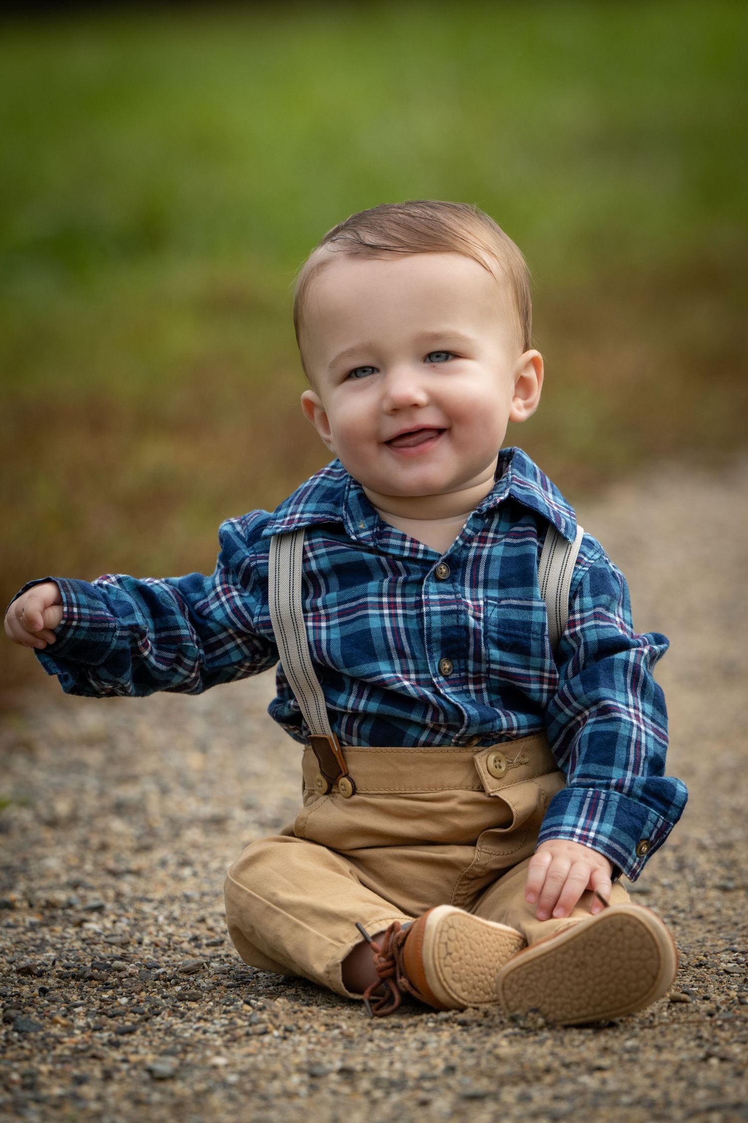 A baby wearing suspenders and plaid shirt sits on a path with a bright smile. Massachusetts Family photographer Serving New England