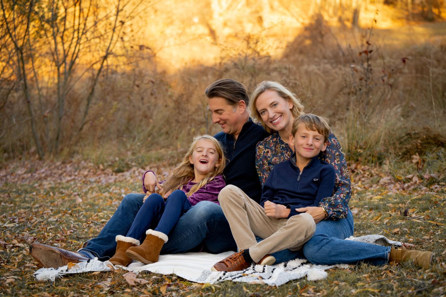 A family sits together on a blanket outdoors during golden hour with autumn foliage in the background.