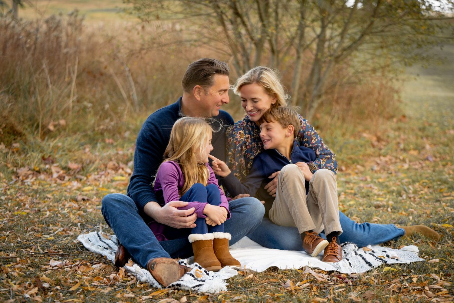 A family enjoys a cozy autumn picnic together on a blanket with fallen leaves surrounding them in nature.