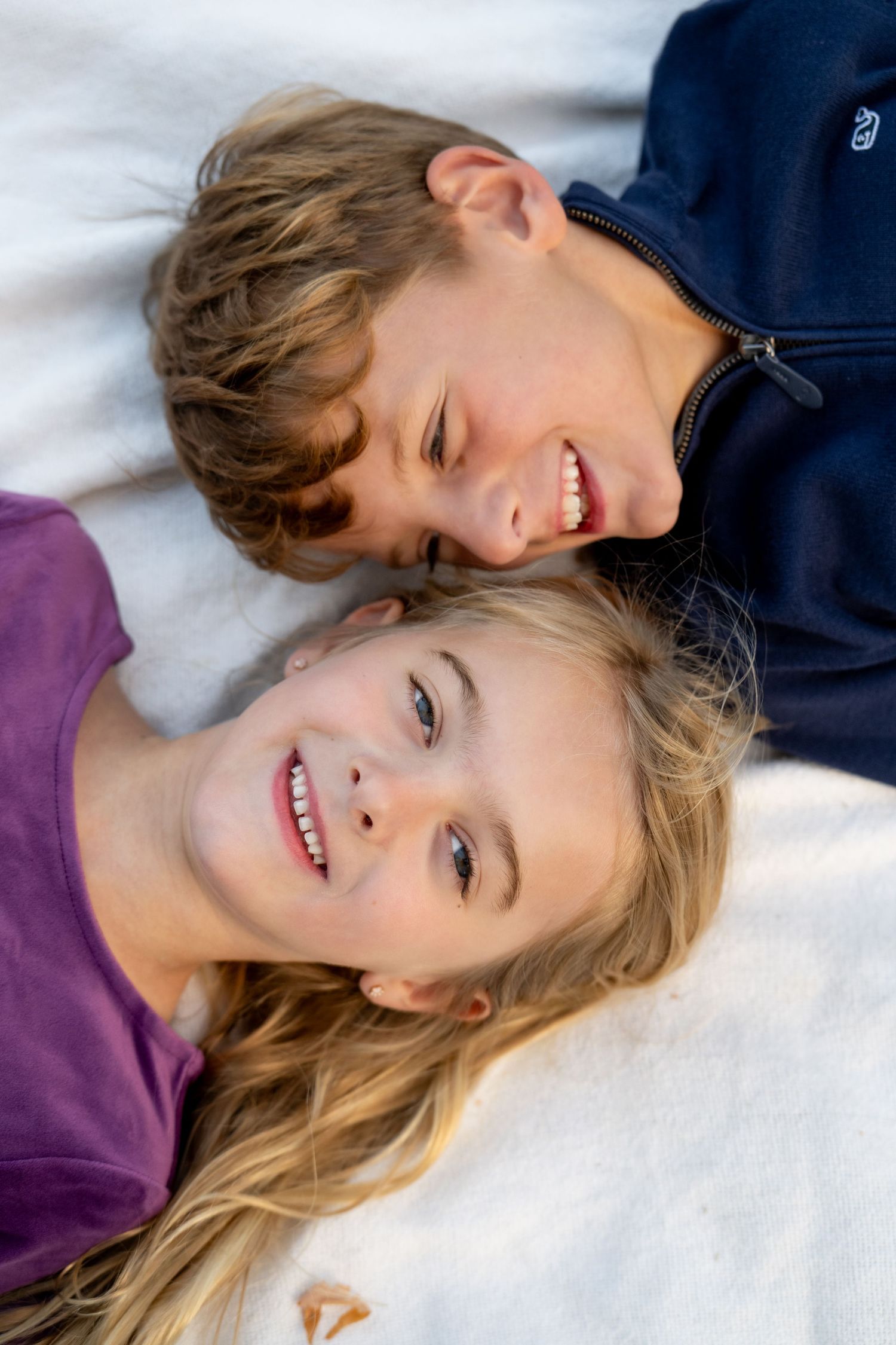 Two children lie on white bedding while smiling and laughing together.