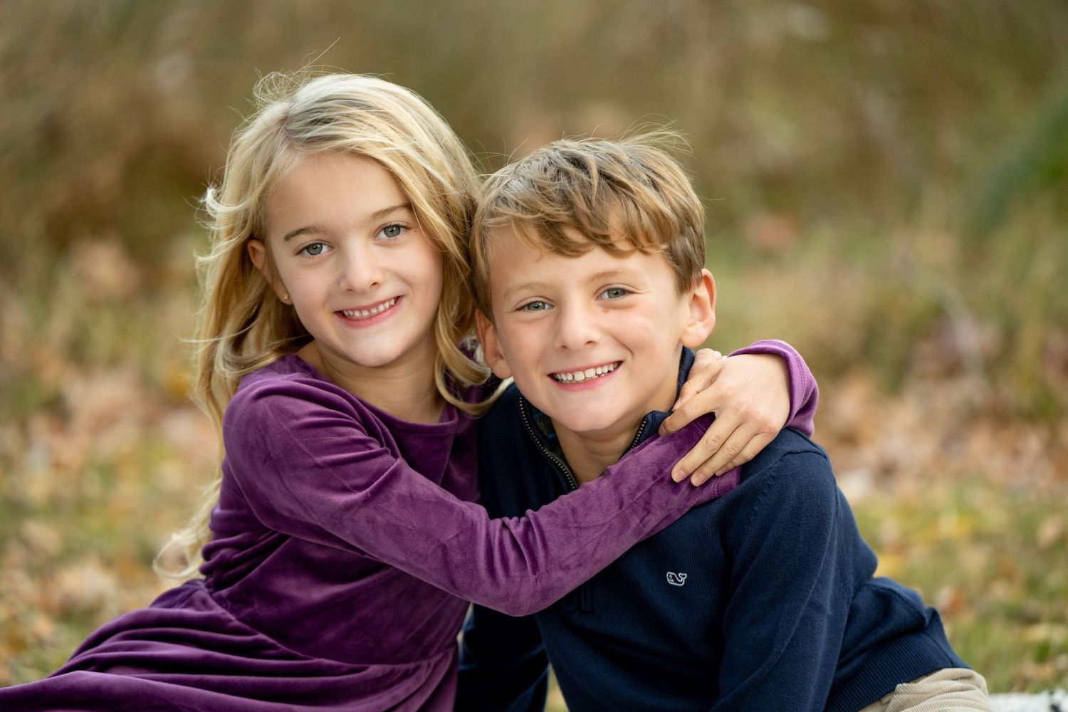 Two children sit close together outdoors smiling, with one wearing a purple dress and the other in navy blue.