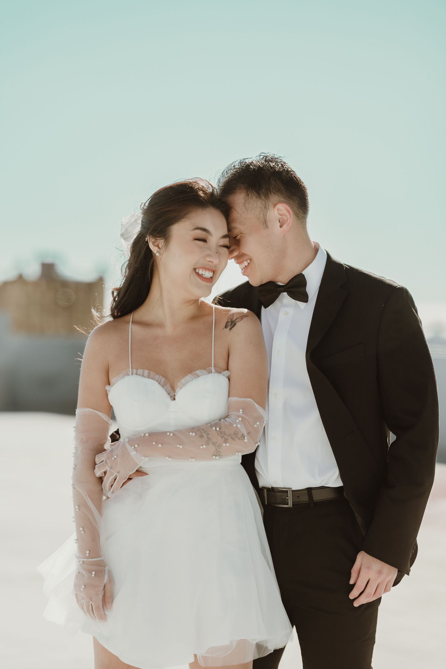 A couple shares an intimate moment in white formal attire against a bright sky.