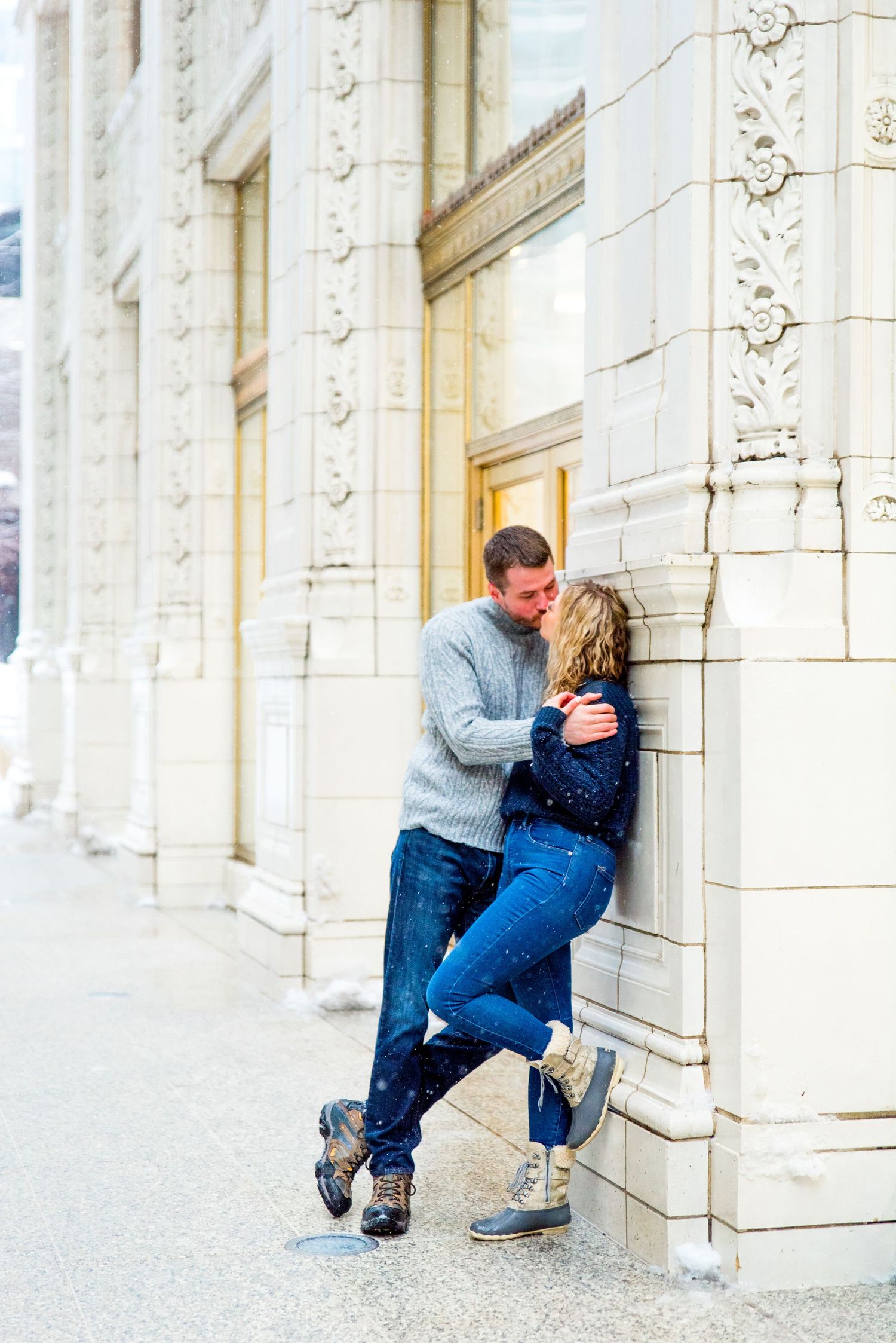 couple in grey and navy sweaters kissing against wall at Wrigley Building in the snow