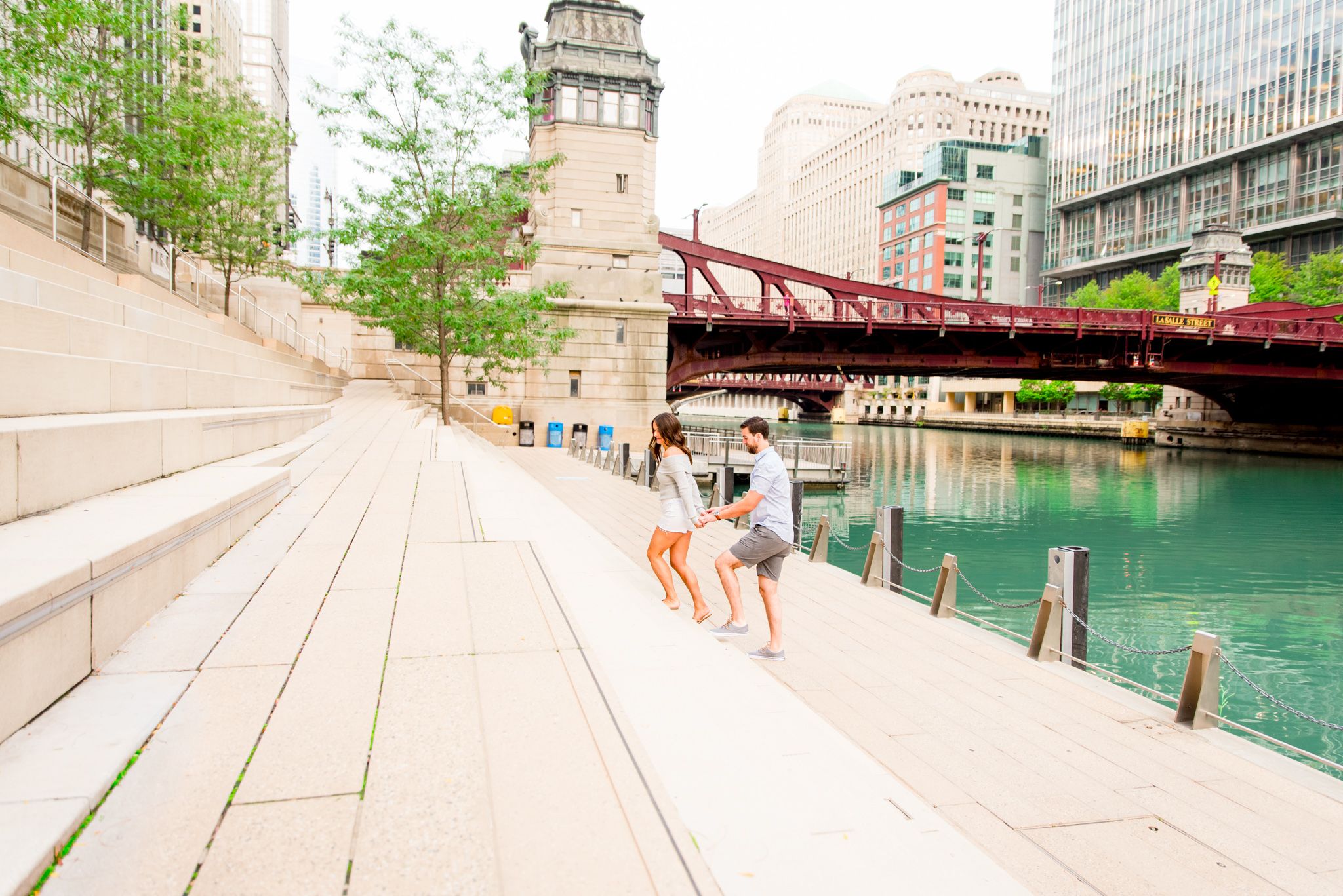 woman leading man up the steps at Chicago Riverwalk for engagement pictures in front of LaSalle Street Bridge