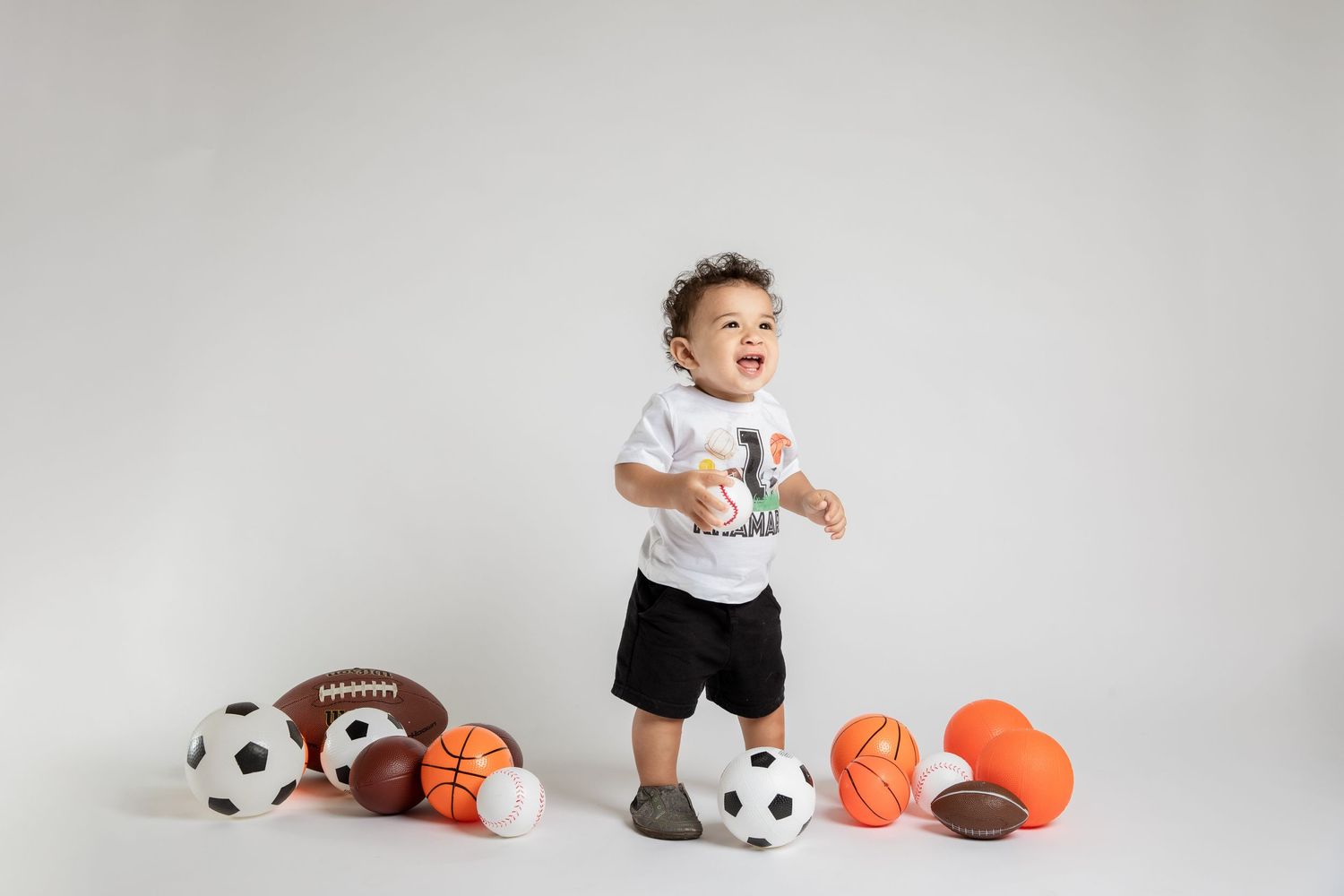 Toddler standing among sports balls including soccer ball, football and basketball.