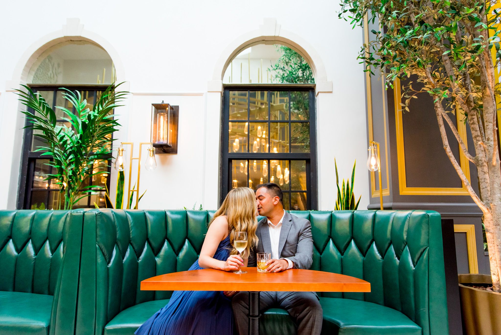 man in grey suit and woman in blue dress holding a glass of white wine kiss in green booth at Lytle Park Hotel