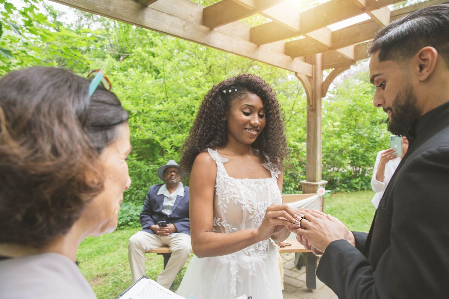 A couple exchanges rings during their outdoor wedding ceremony under a wooden pergola.