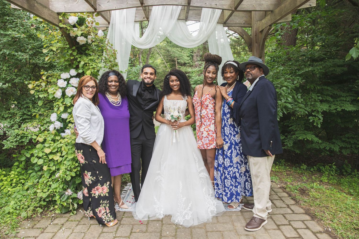 A wedding party poses together under a decorated outdoor archway in a garden setting.