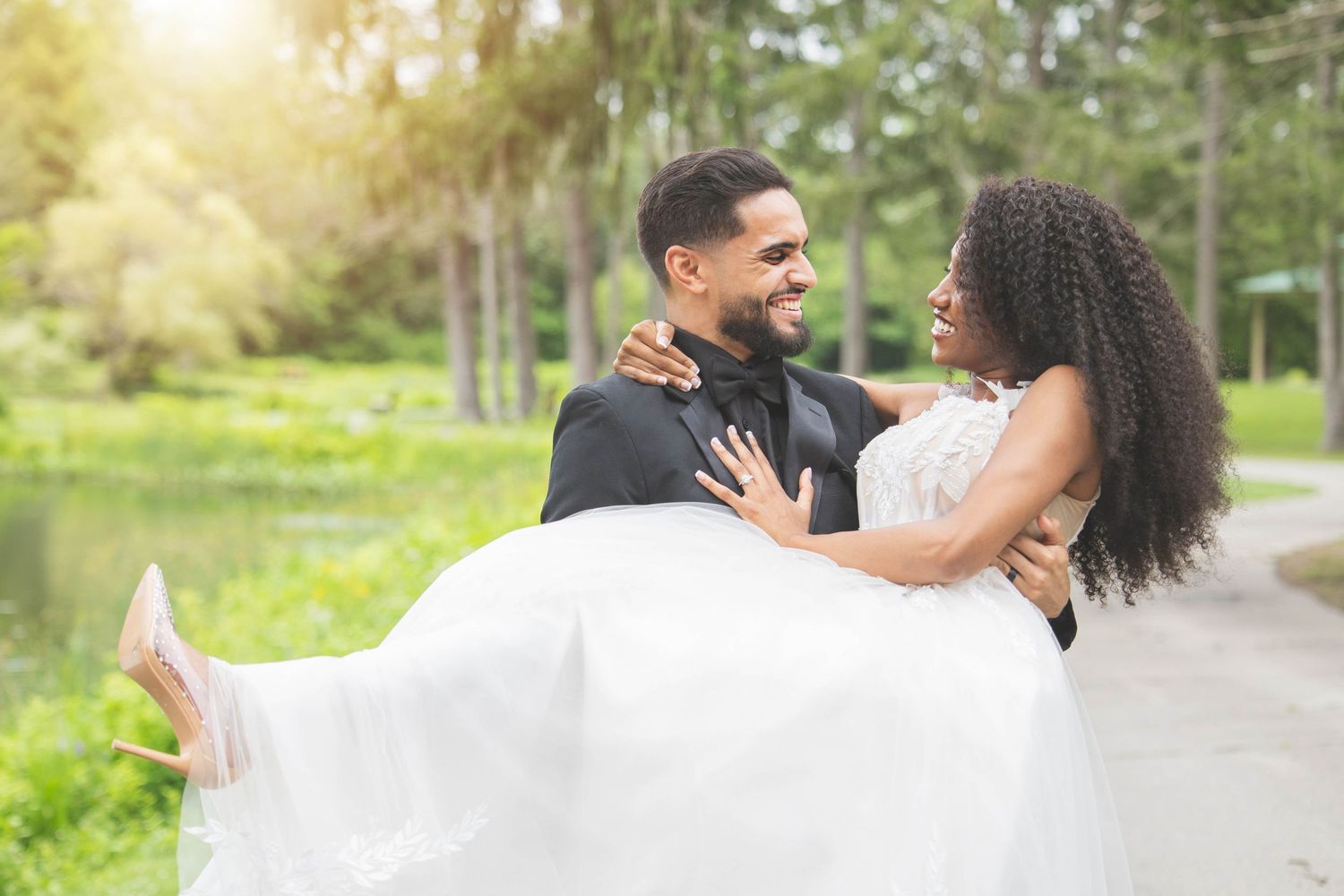 A romantic wedding photo of a groom carrying his bride in a white dress on a sun-lit path.
