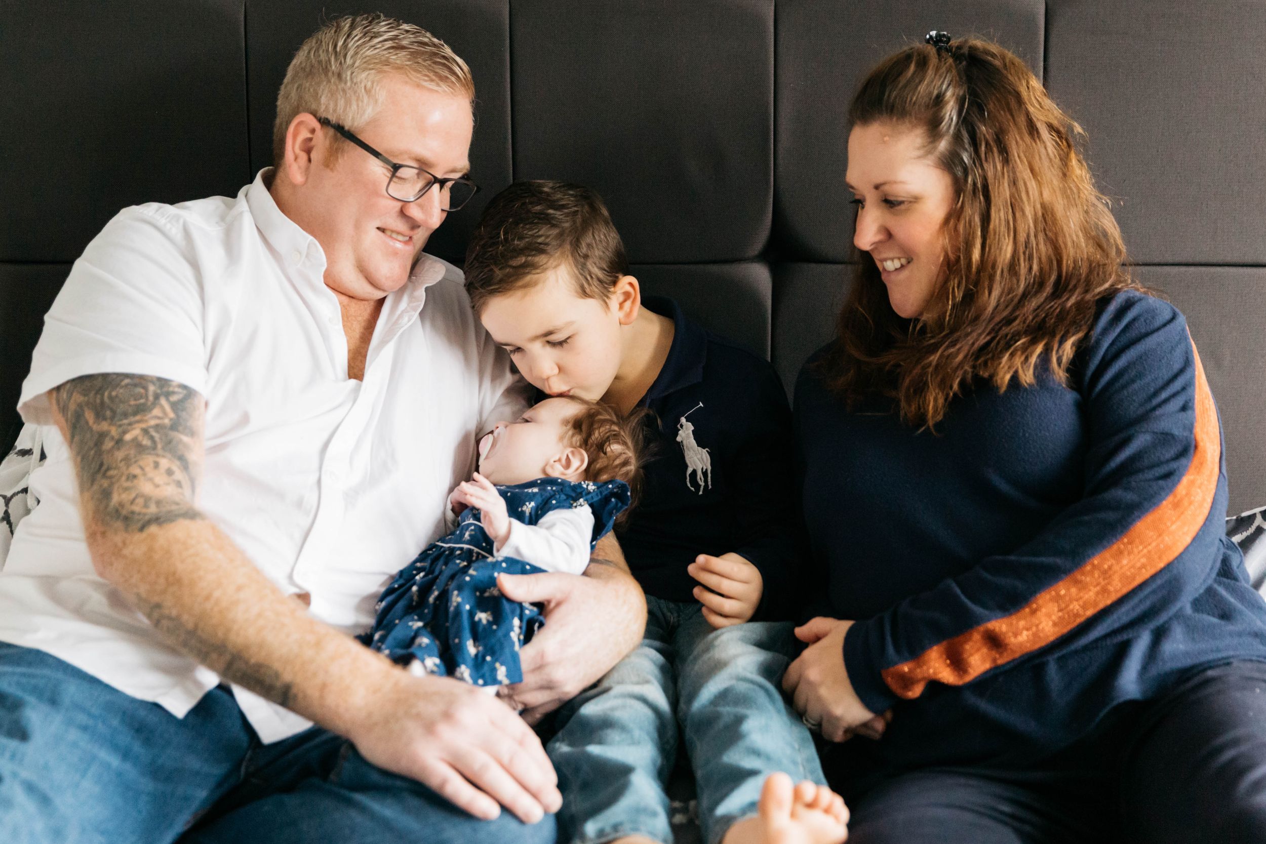 family of four sitting on bed big brother kissing baby sister head