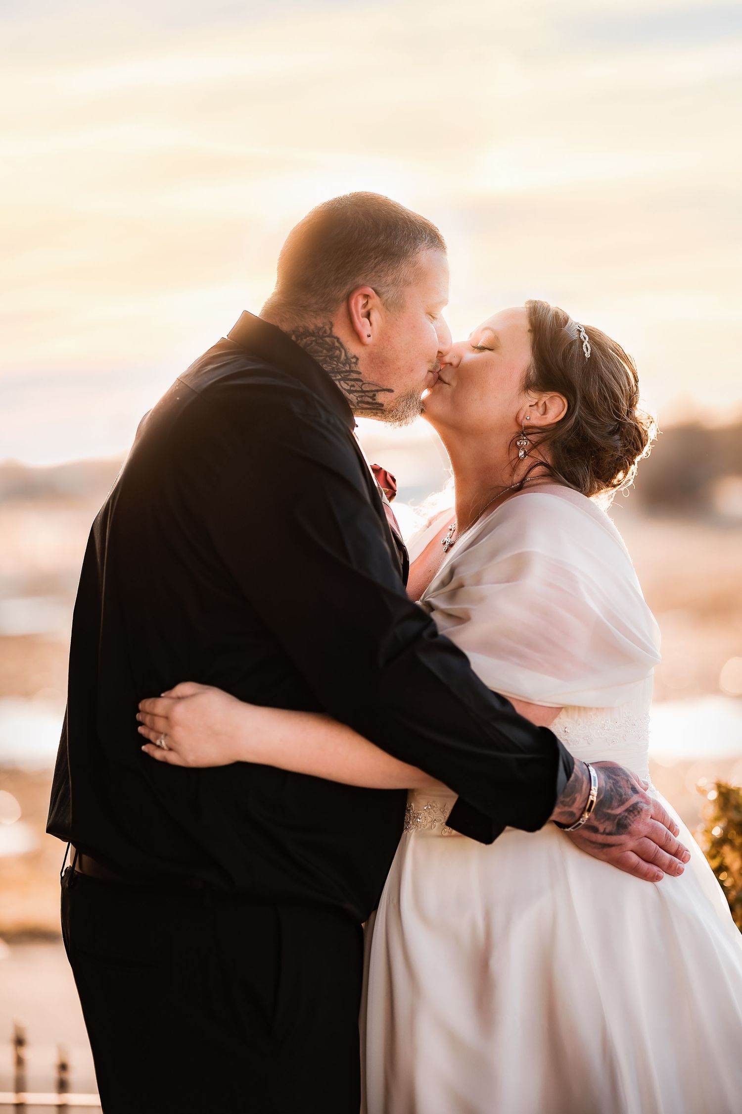 A couple shares a romantic kiss at sunset while wearing formal wedding attire.