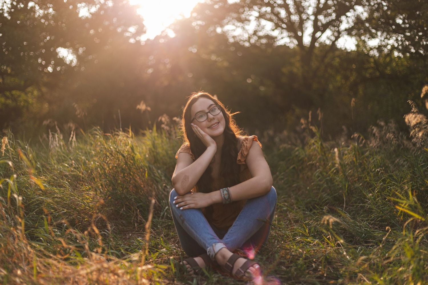 A senior sitting cross-legged in a field of tall grass with the sun streaming through the trees in St. Cloud, MN