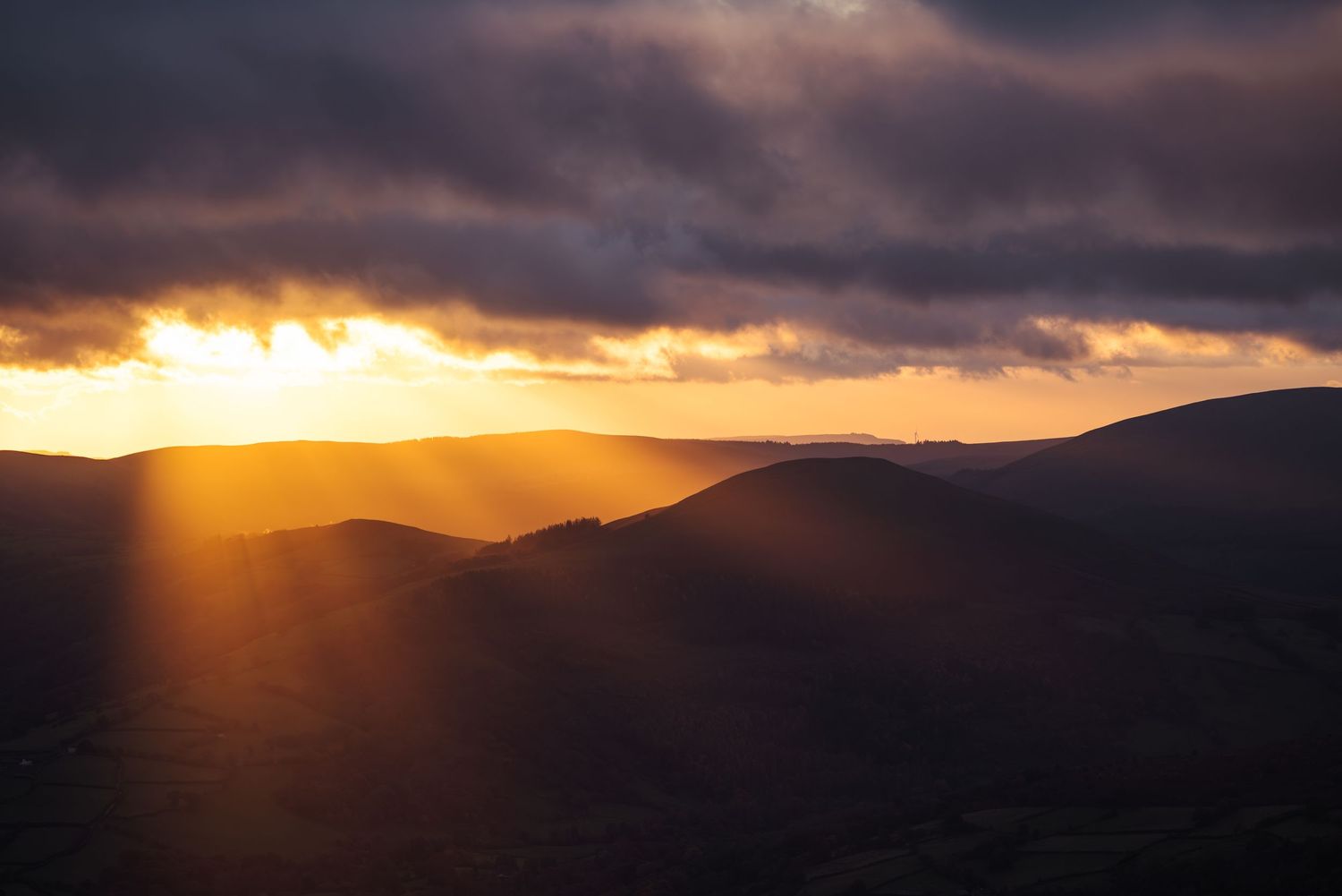 Dramatic sunset rays pierce through dark storm clouds over mountainous terrain.