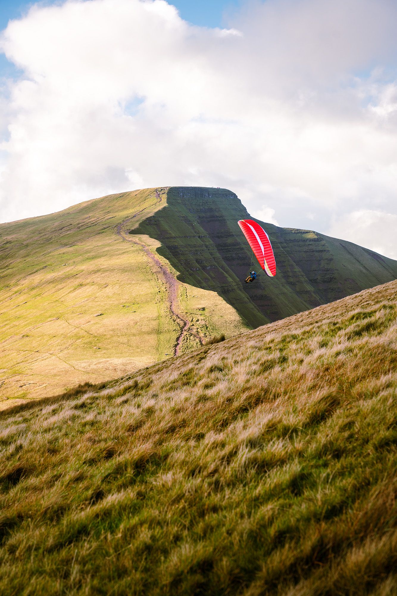 Red paragliding wing floats against a steep grassy mountainside with hiking trails visible.