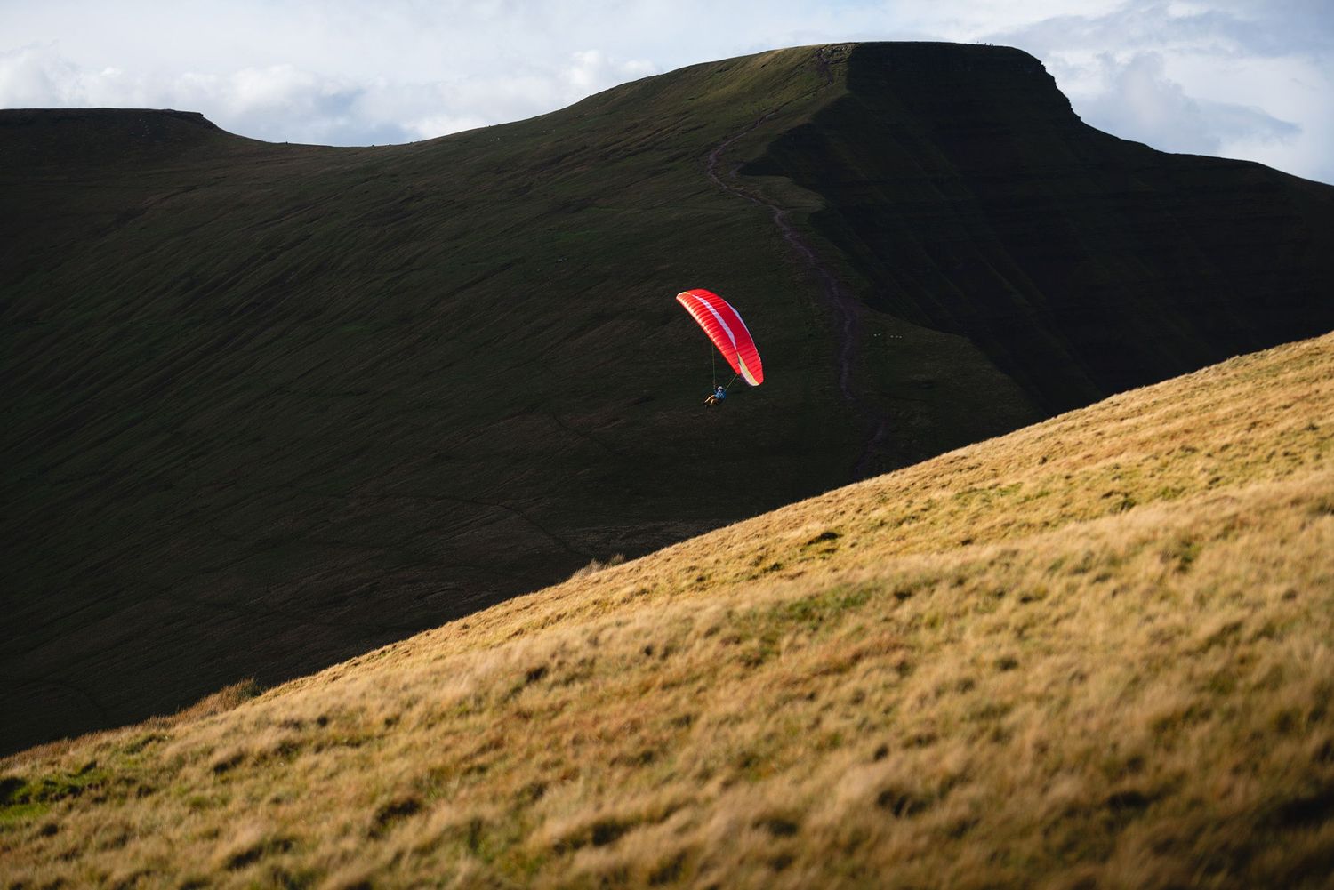 A red paragliding wing glides against a dark mountain backdrop with grassy slopes.