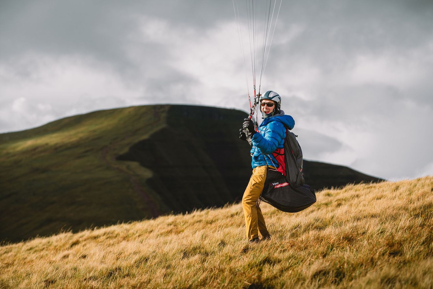 A paraglider in blue and yellow gear soars above grassy mountain slopes.