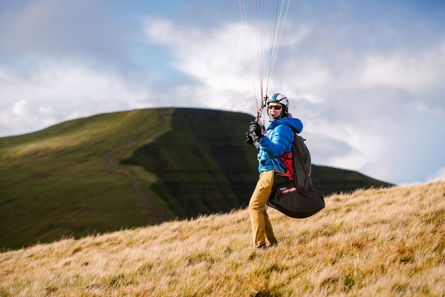 Paraglider soaring over golden hillside against cloudy sky in blue and tan gear.
