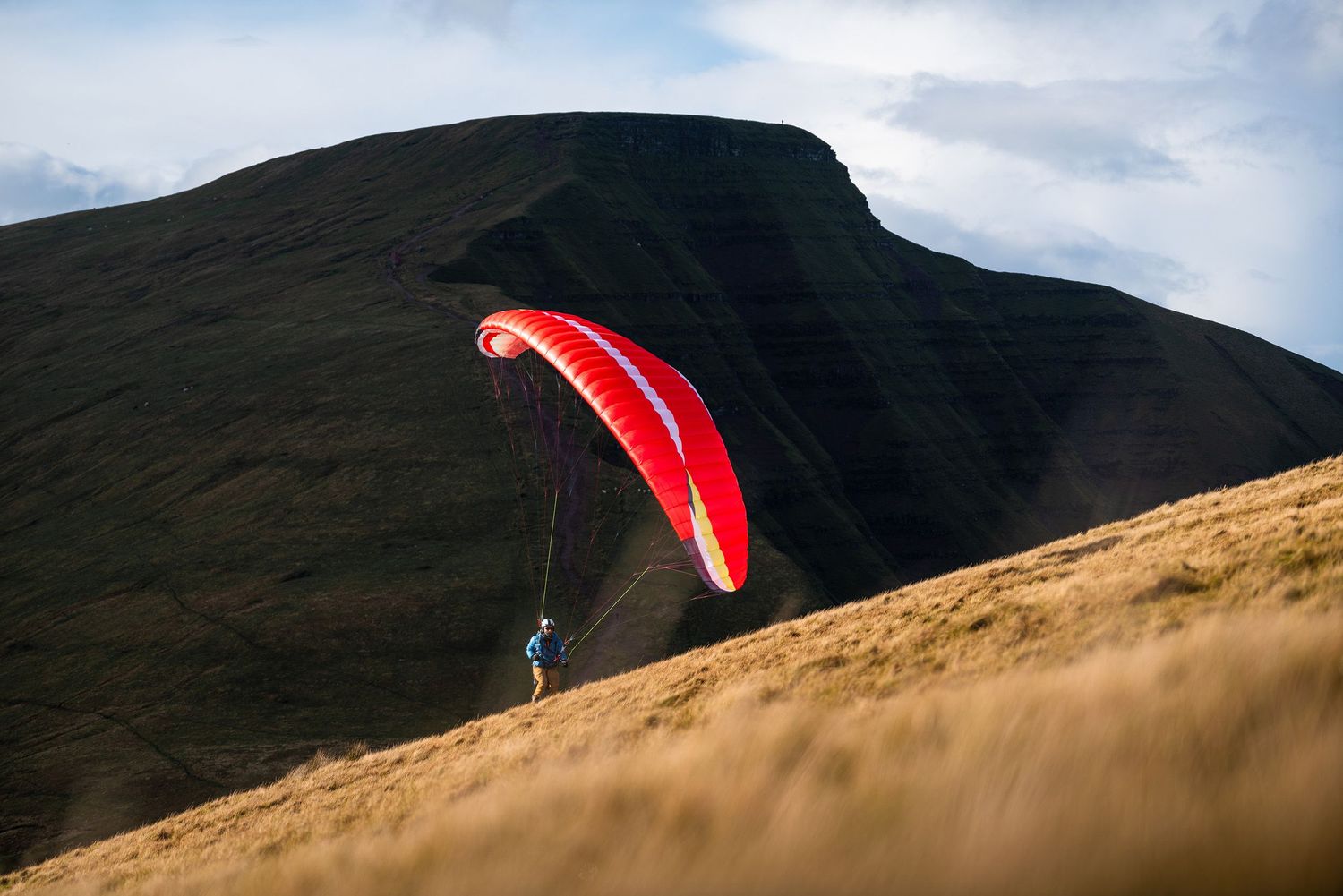 Bright red paraglider wing glides across steep mountain terrain under cloudy skies.