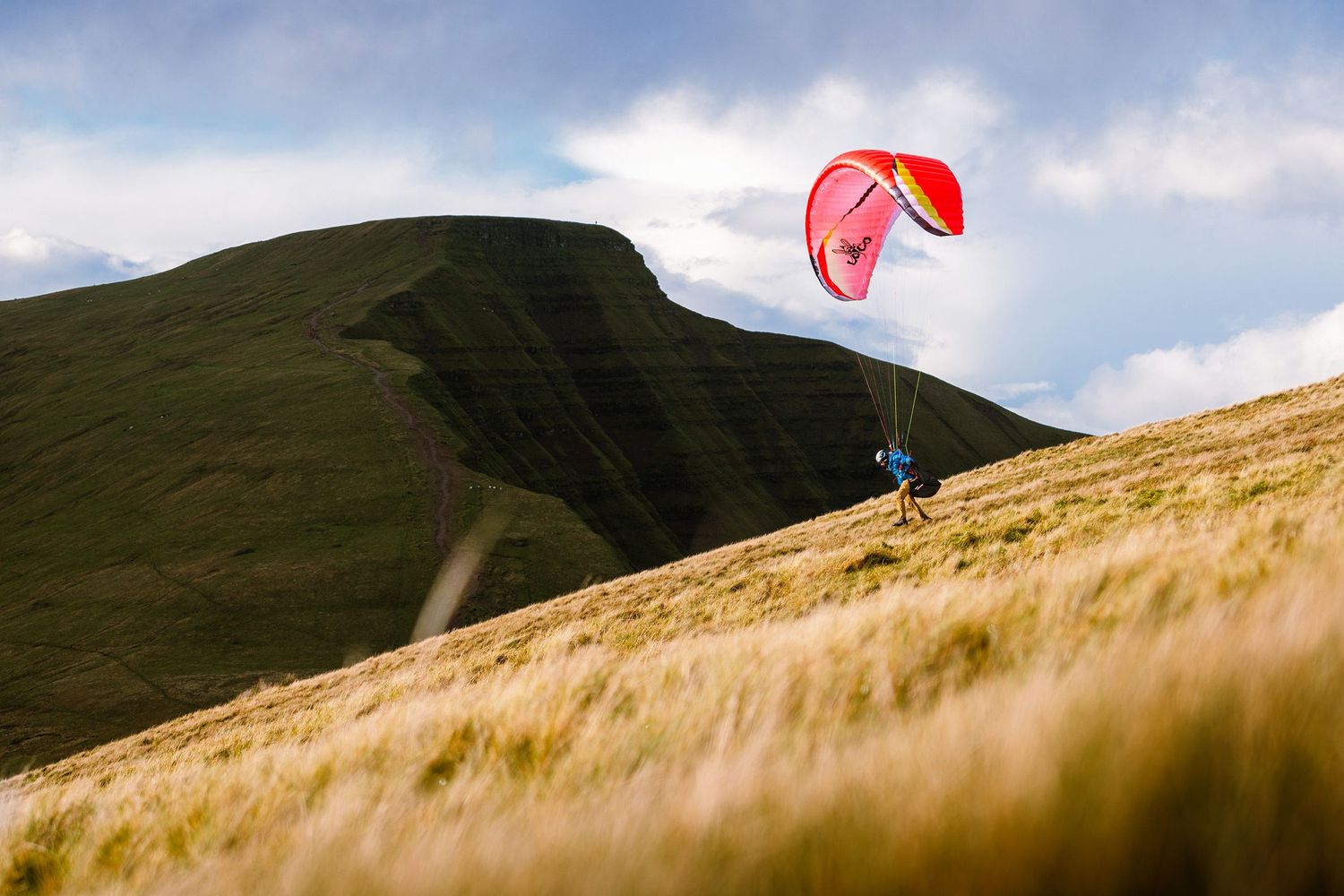 Red paragliding wing soars above golden grass-covered mountain slopes.