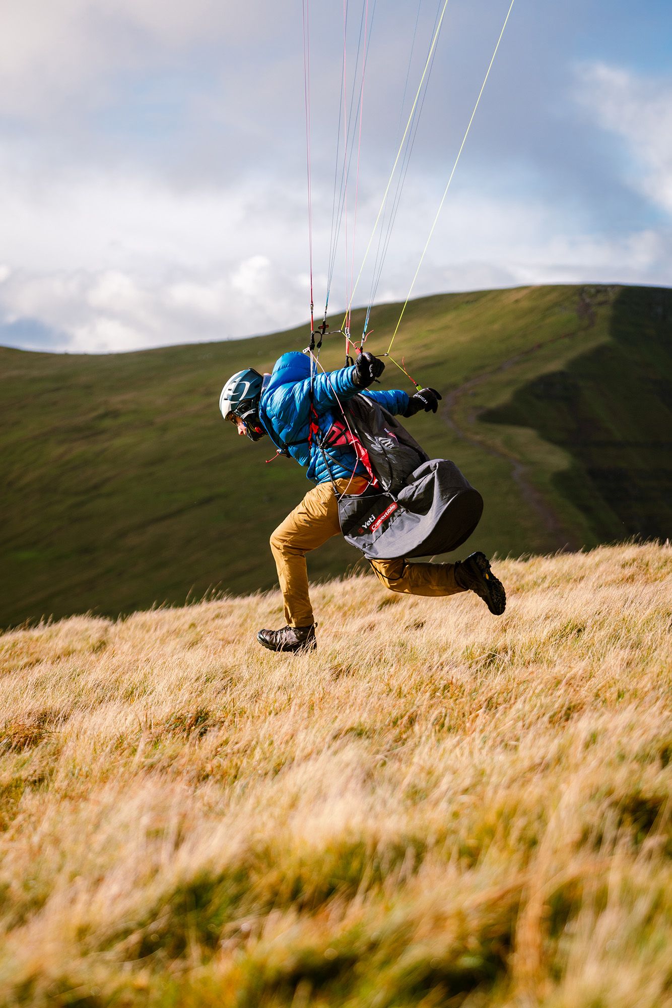 A paraglider in motion wearing blue jacket and yellow pants against grassy hillside.