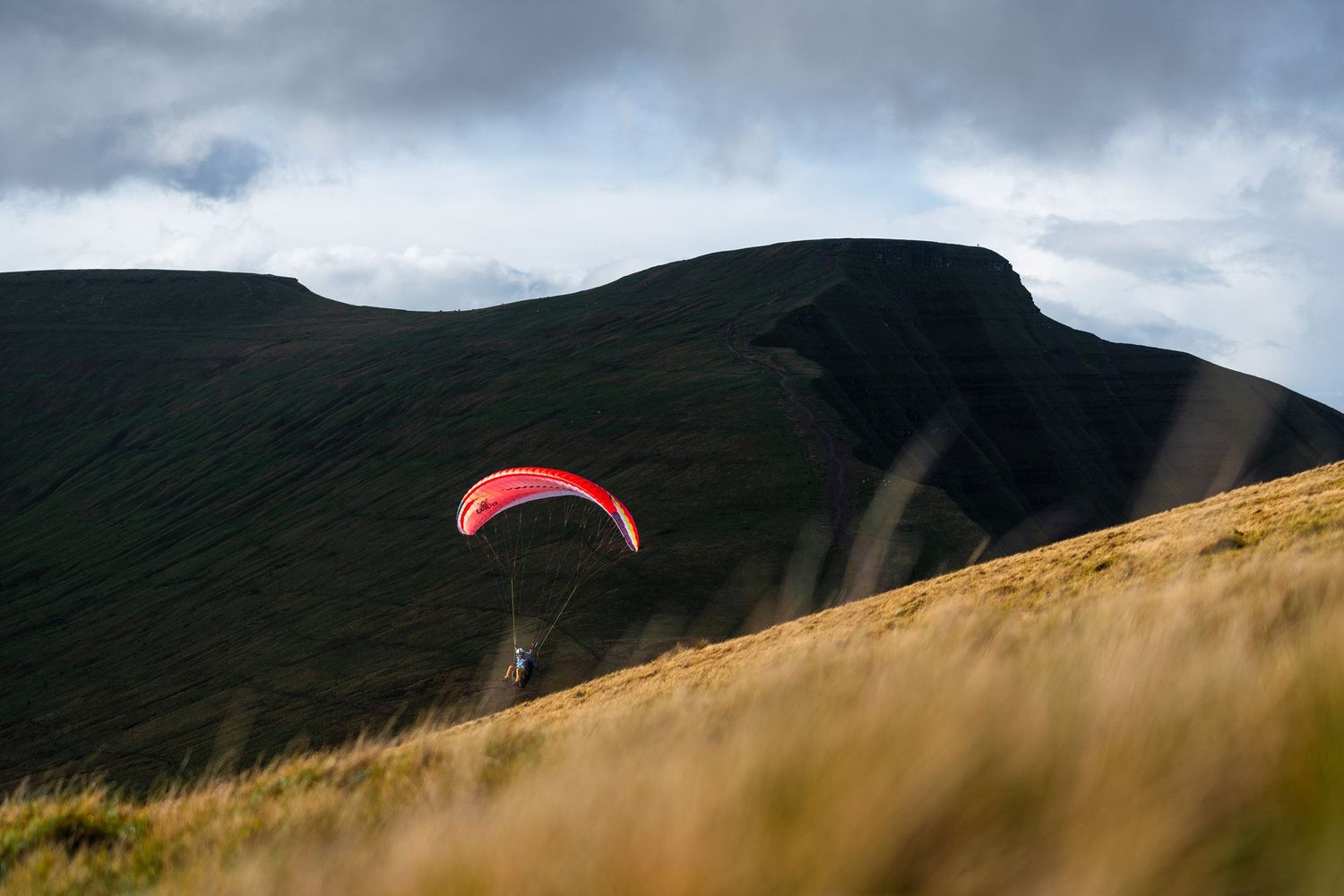 A red paraglider wing contrasts against dark mountain slopes and moody sky.