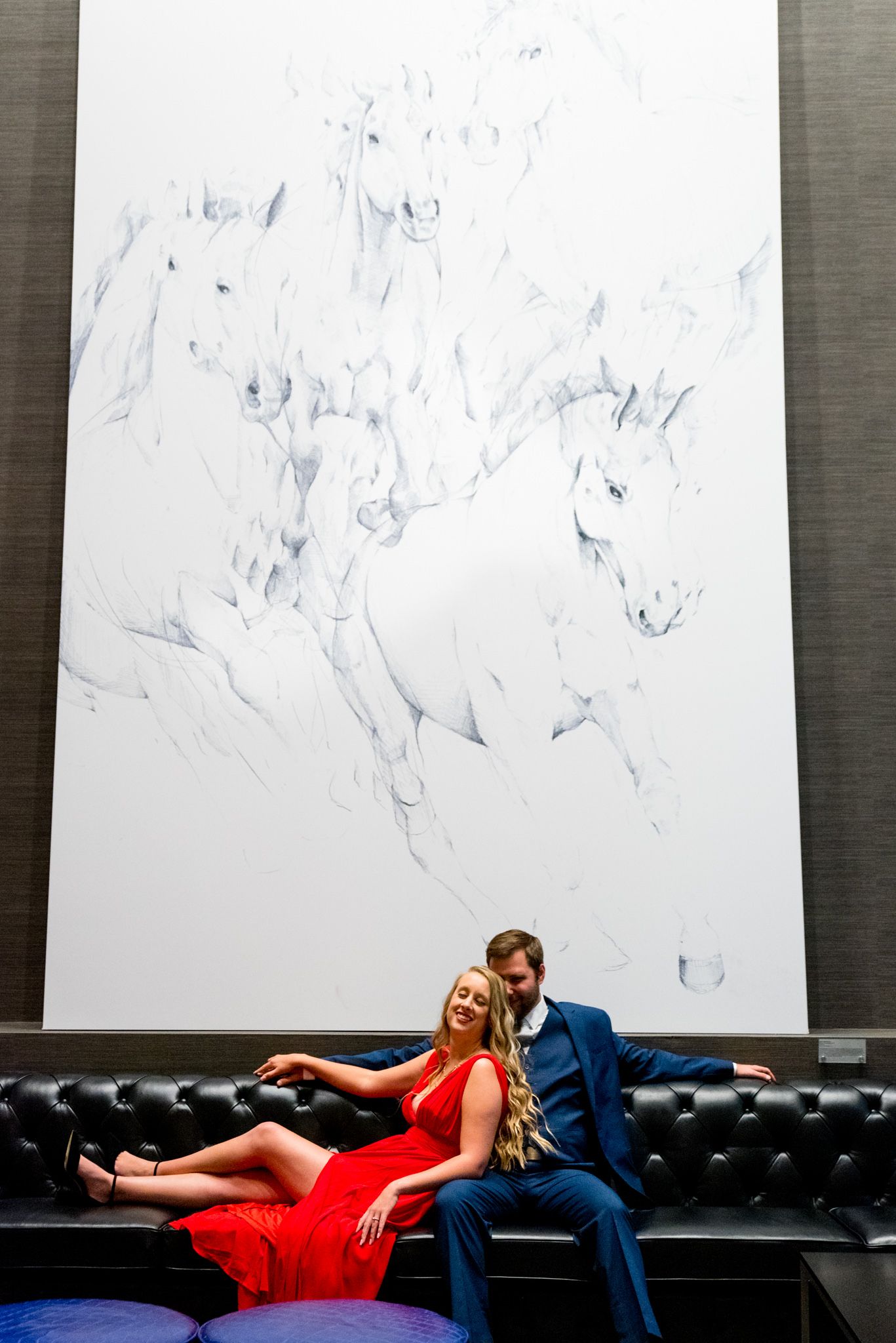 man in navy suit sitting on black leather couch and woman in red dress leans back and laughs in front of horse mural