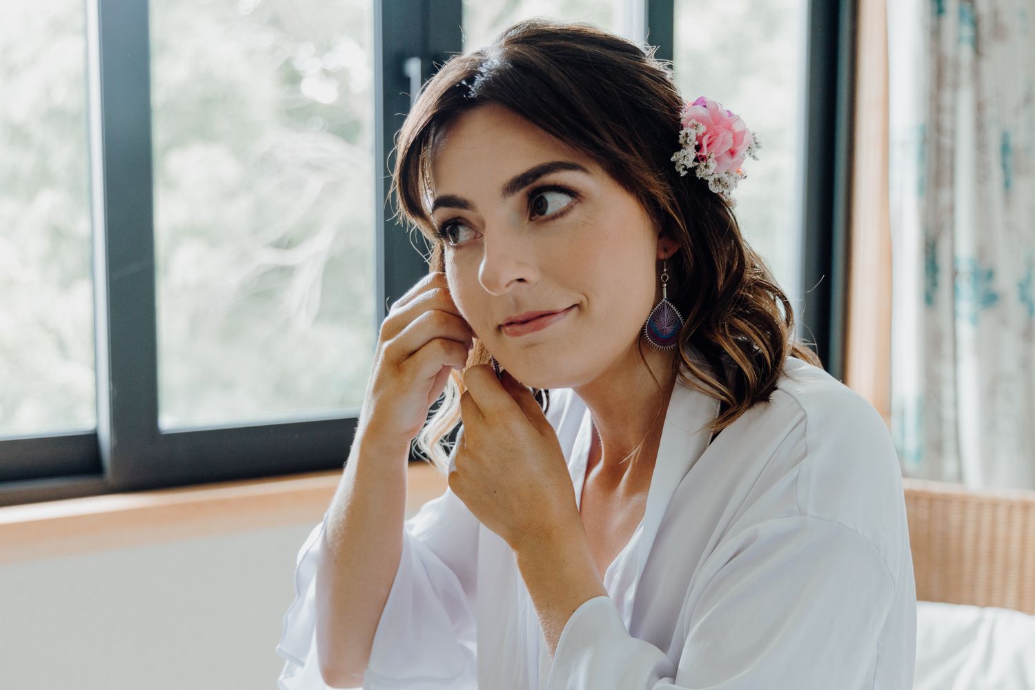 A person in a white robe with pink flowers in their hair gazes thoughtfully to the side in a bright room.
