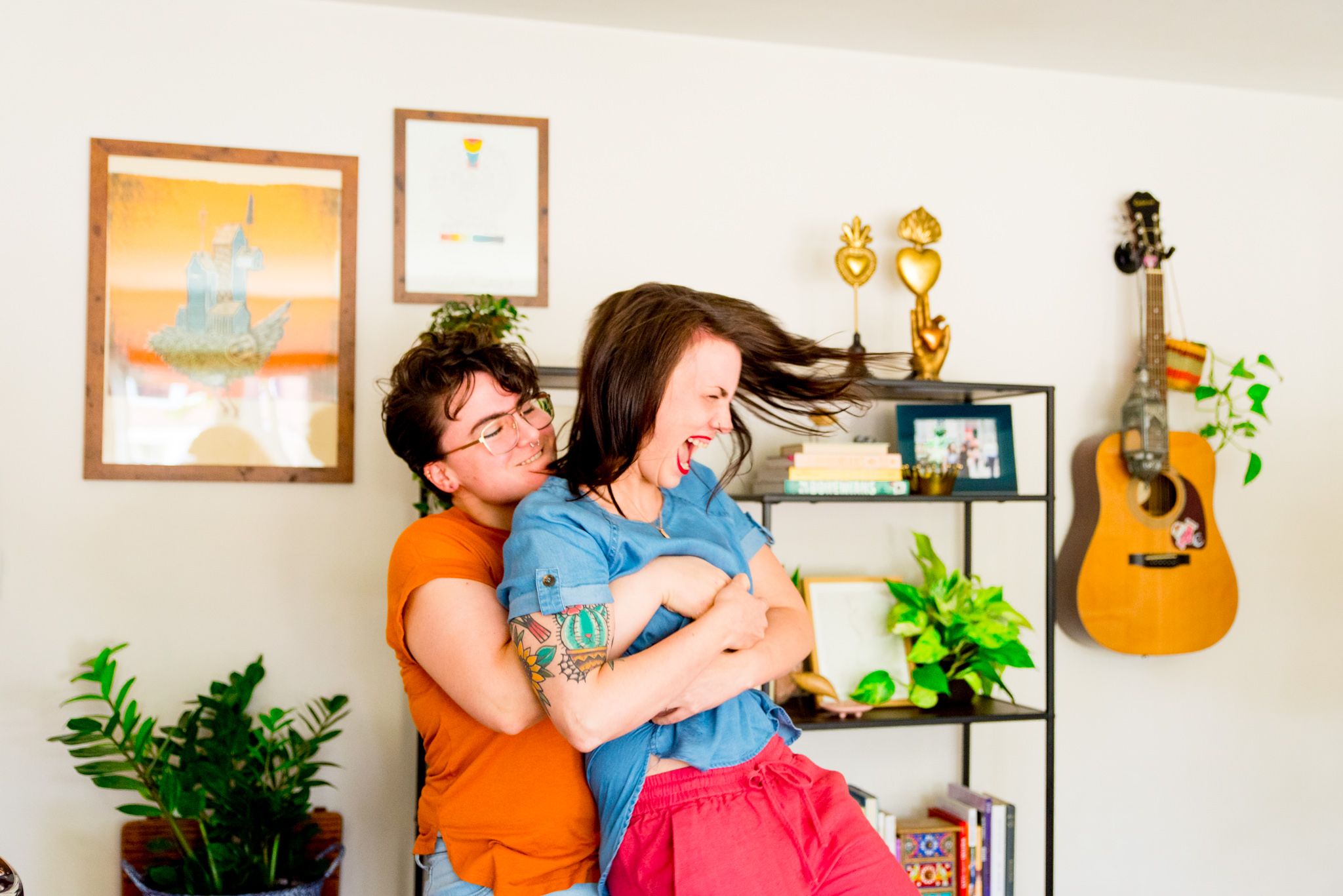 woman in orange smiling picking up wife in pink pants laughing and flipping her hair in front of wall with guitar hangin