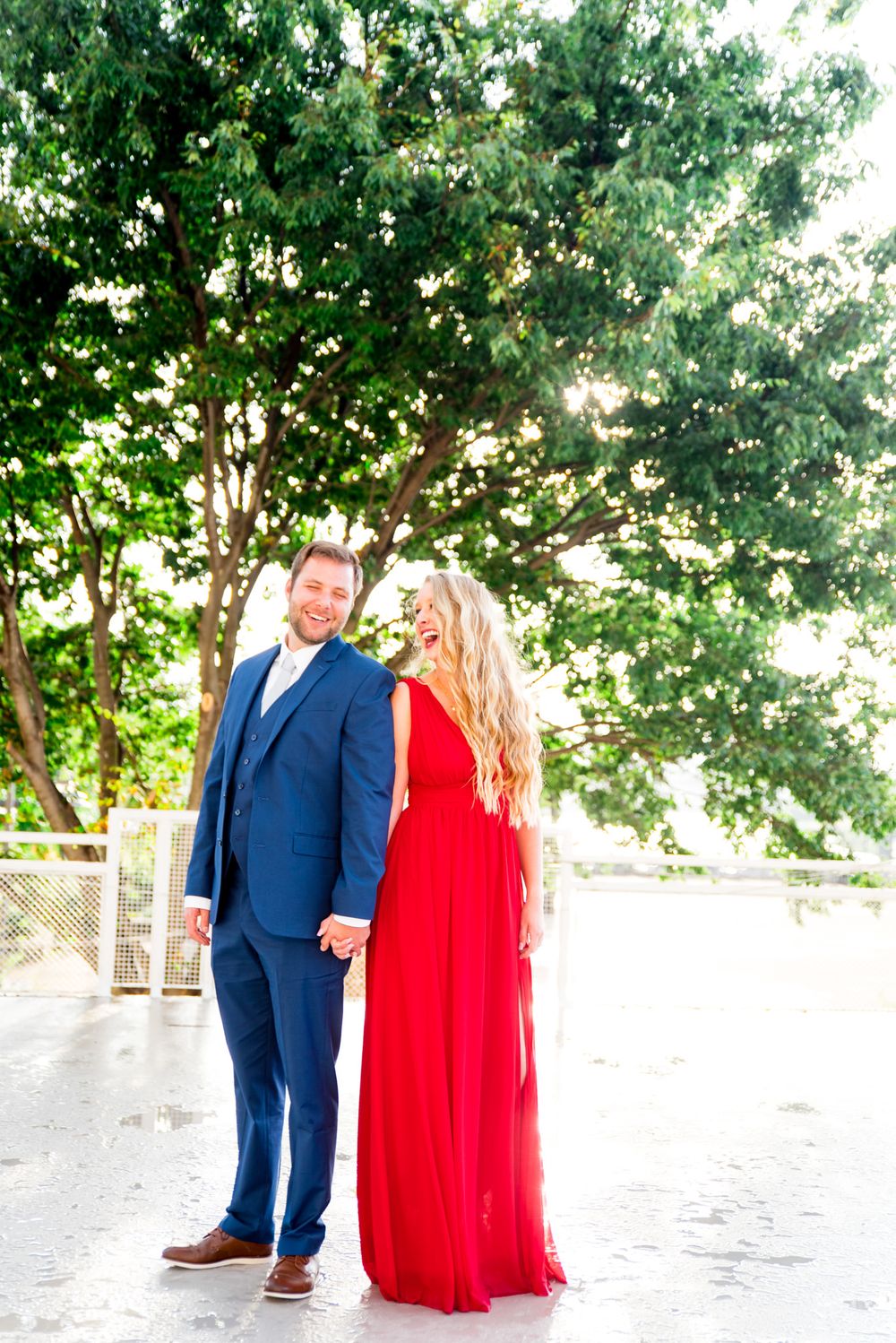 blonde woman in red dress and man in navy suit look at each other laughing in front of big tree
