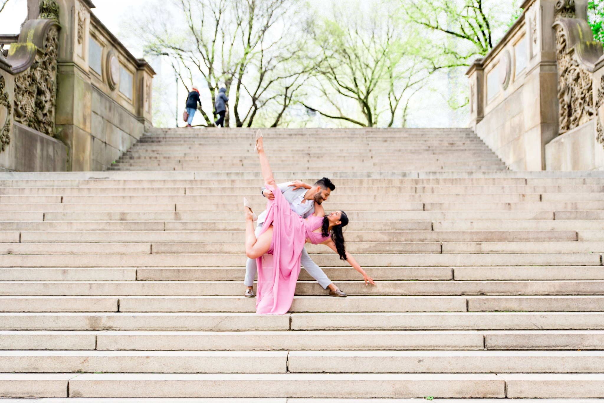 Woman in pink dress and man in gray suit dancing on steps to Bethesda Terrace Central Park for spring engagement photos