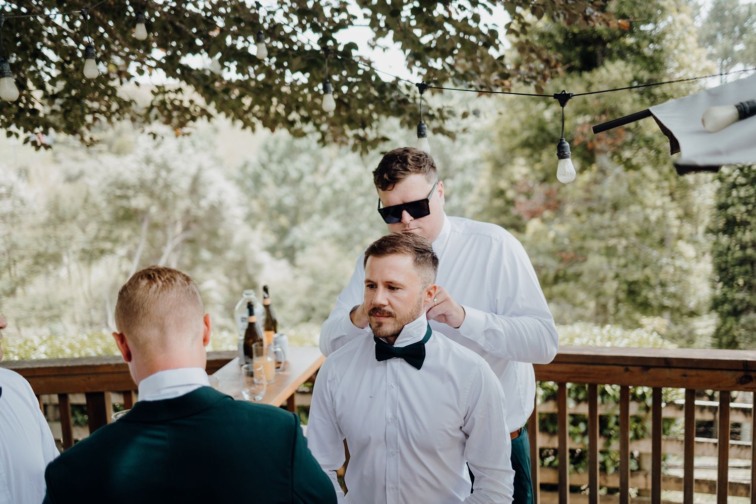 People in formal attire enjoying drinks on an outdoor deck surrounded by trees.