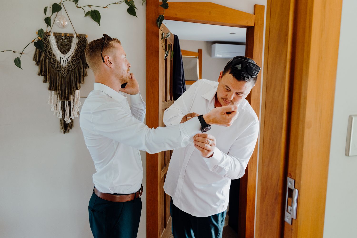 Two people in white shirts help each other get dressed in a room with wooden decor.