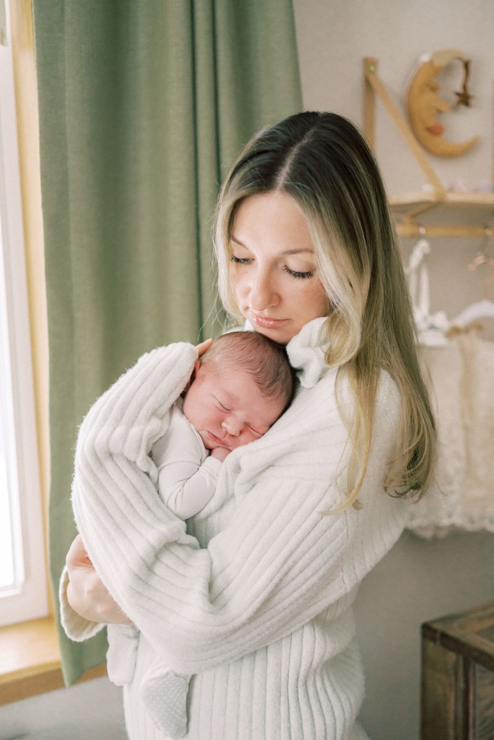 Tender moment with newborn sleeping against white sweater near window.