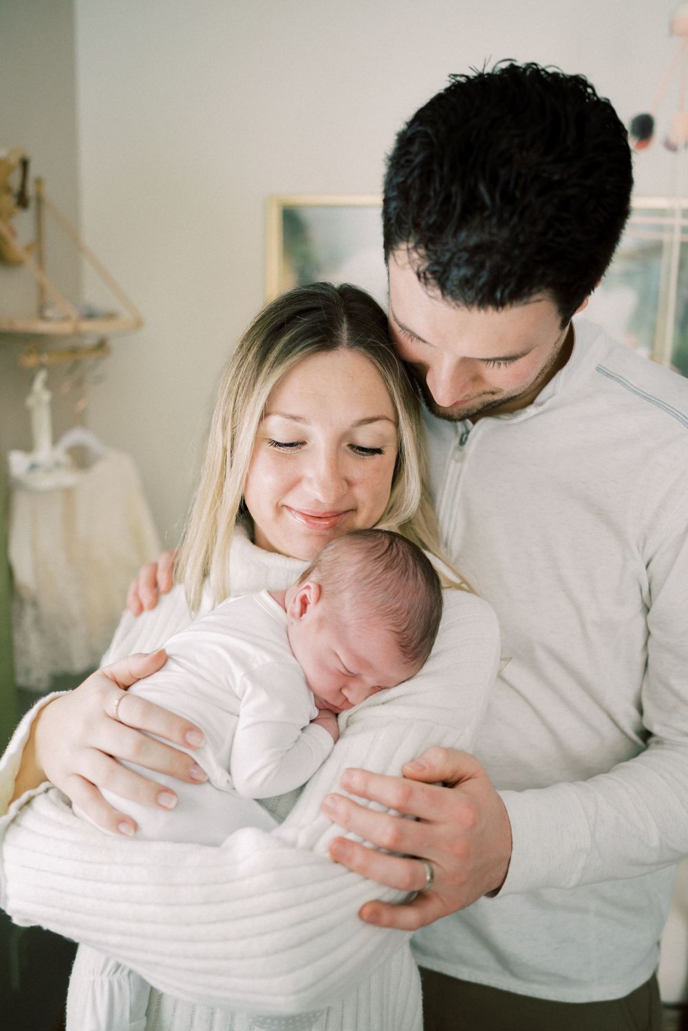 Family moment as parents in white sweaters embrace their sleeping newborn.