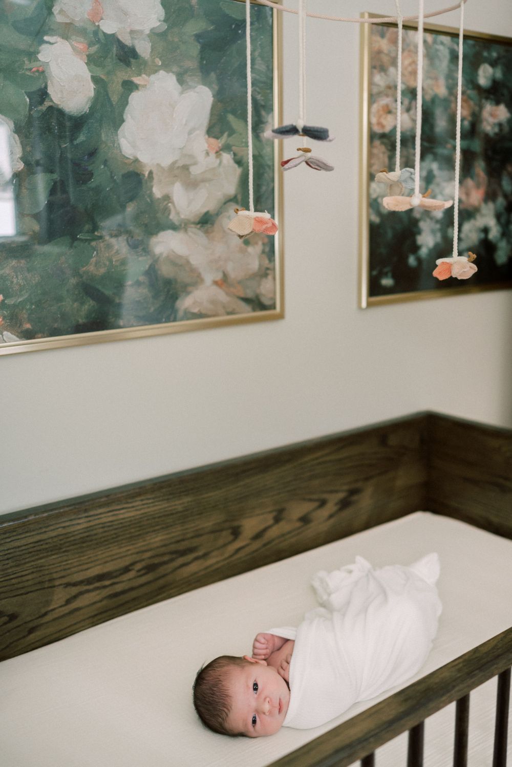 Newborn lying in wooden crib beneath floral artwork and hanging decorations.