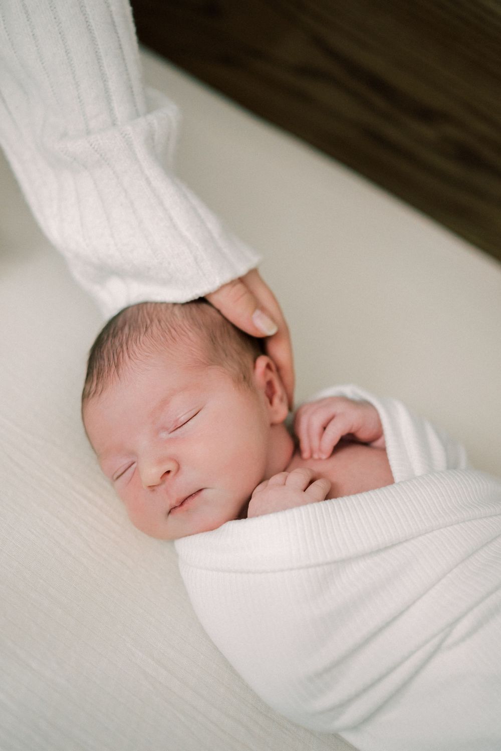 Sleeping newborn baby swaddled in white fabric while being gently touched.