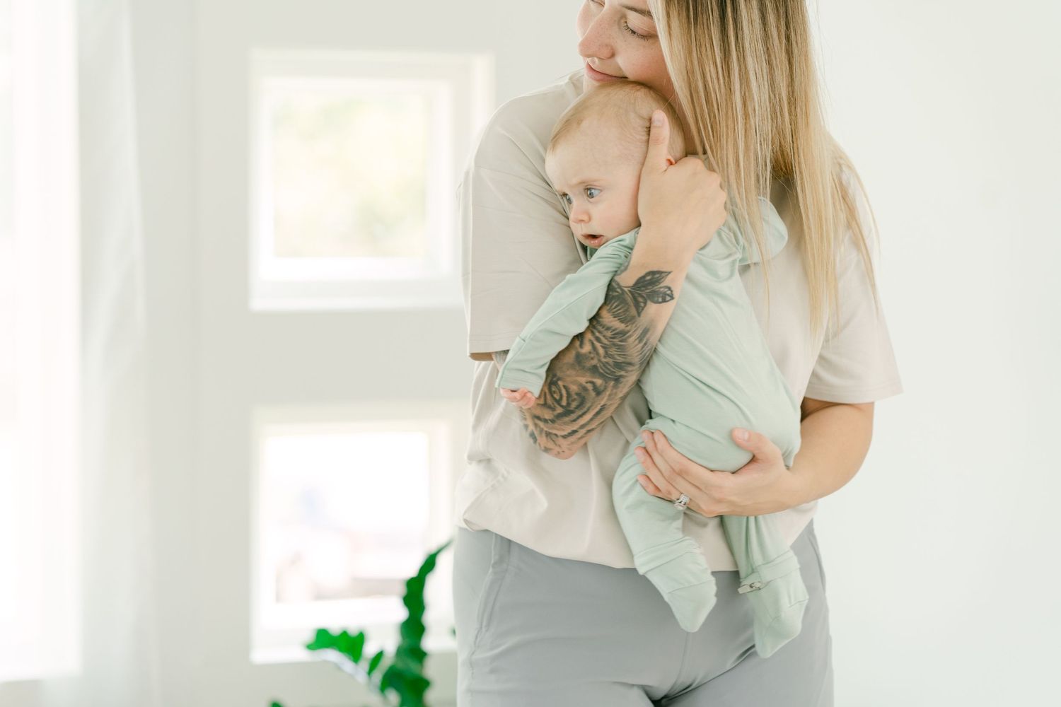 Parent in casual clothes holding baby in soft mint green outfit against white wall.