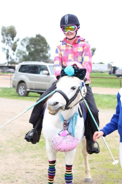 Rider in pink plaid jacket on decorated white pony wearing colorful leg warmers and festive accessories.