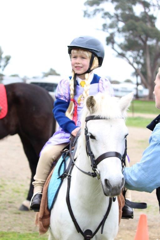 Young rider in blue helmet and colorful outfit sitting atop white pony during equestrian event.