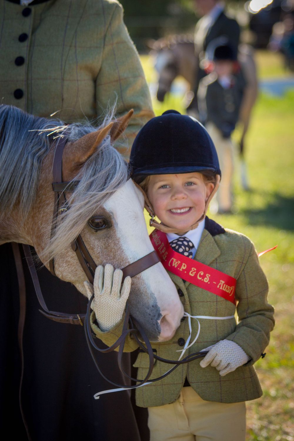 A young equestrian in show attire stands beside a gray horse at an outdoor competition.