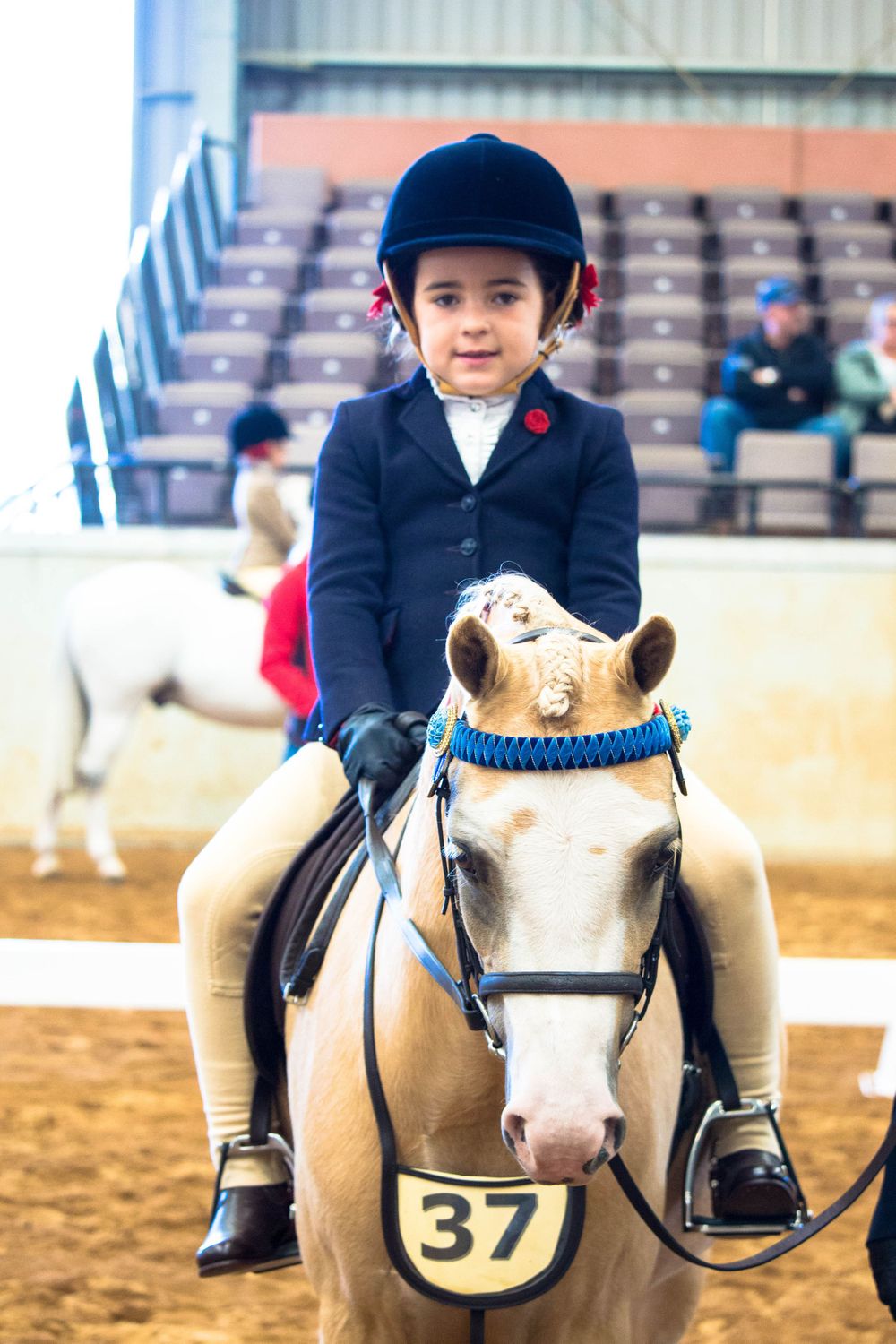 Young equestrian in formal show attire riding a cream-colored pony with competition number.