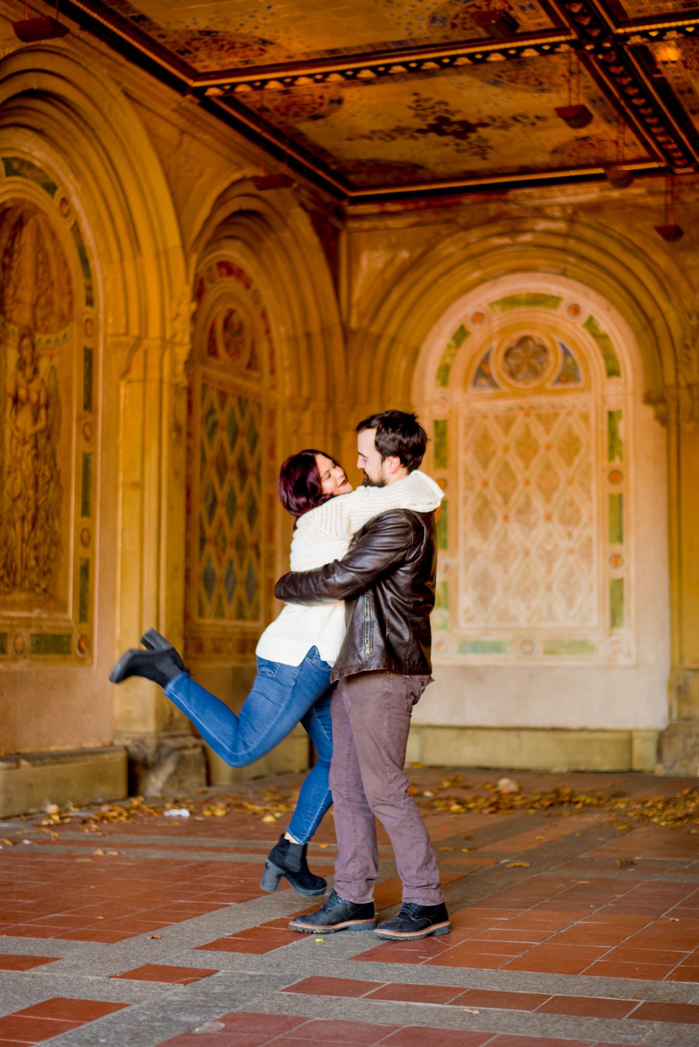 young woman hugging man and kicking feet up at Bethesda Terrace in Central Park for engagement pictures