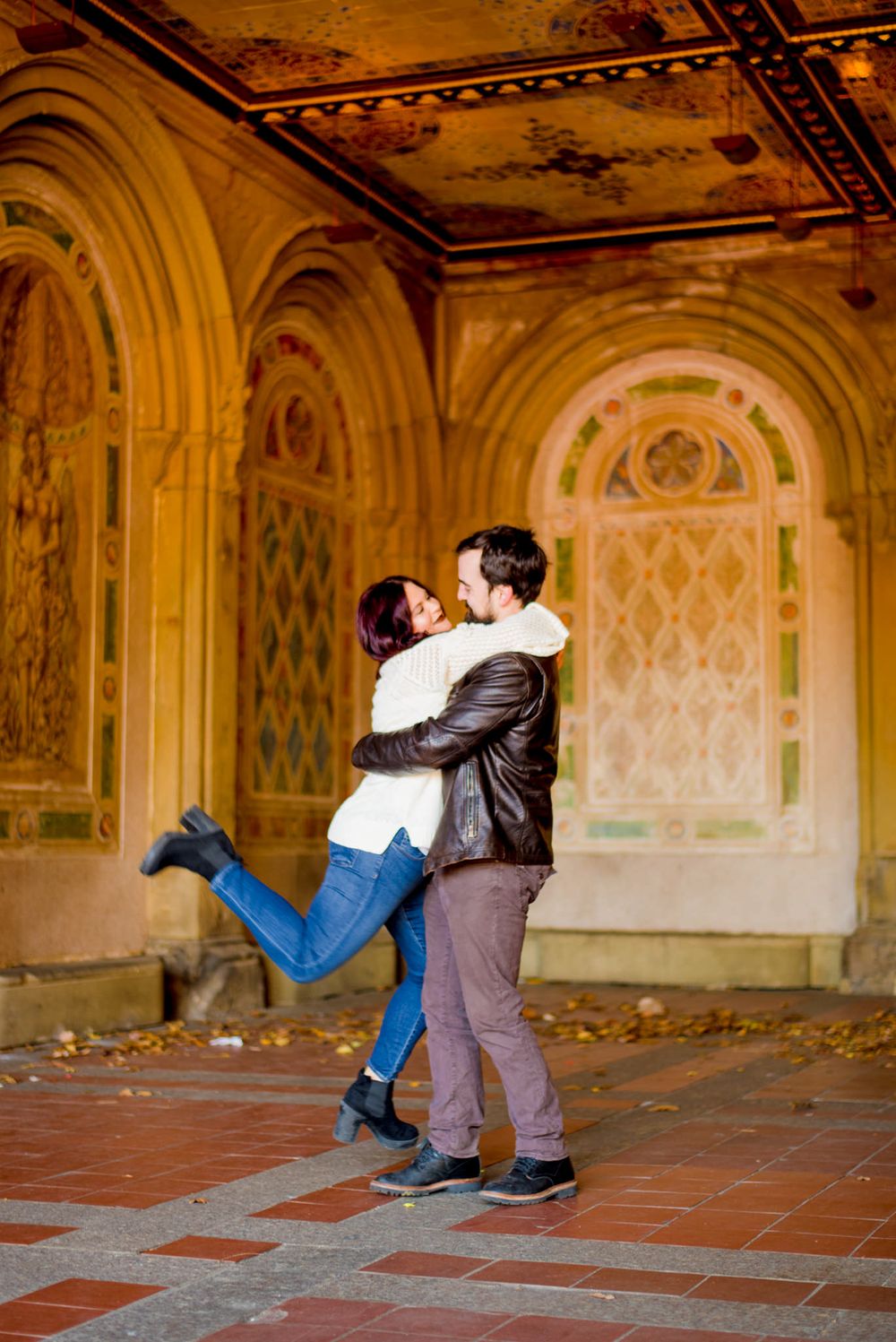 young woman hugging man and kicking feet up at Bethesda Terrace in Central Park for engagement pictures