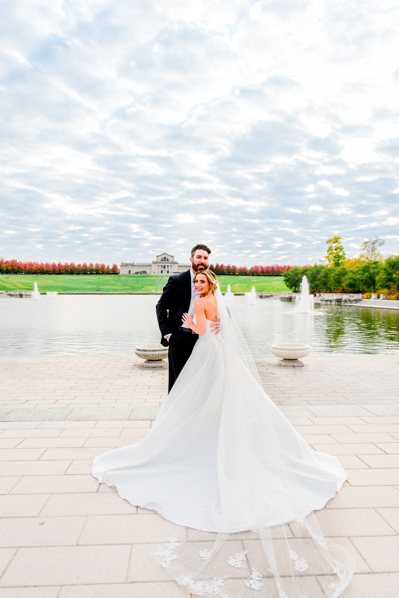 bride's wedding dress and veil fanned out next to groom under blue sky with clouds for Forest Park STL fall wedding