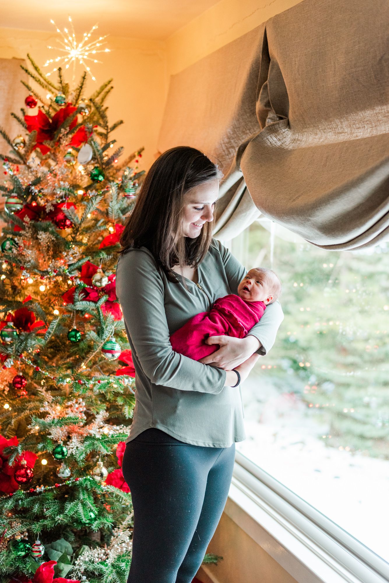 Lifestyle mother holding newborn baby in pink wrap in front of Christmas tree in Pittsburgh, Pennsylvania