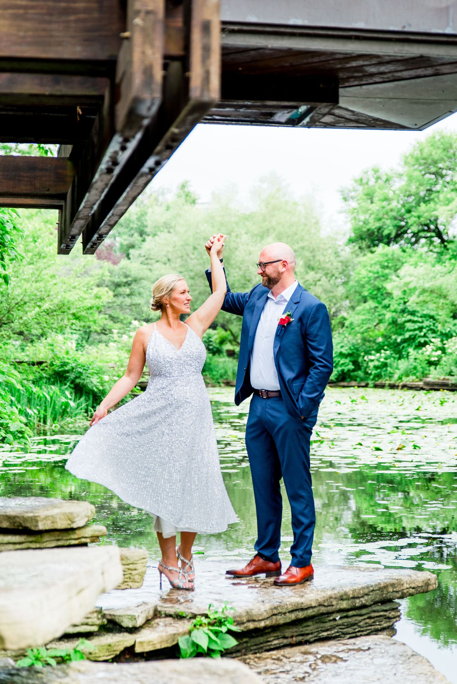 man in navy suit twirling blond woman in silver dress at Alfred Caldwell Lily Pool in Lincoln Park